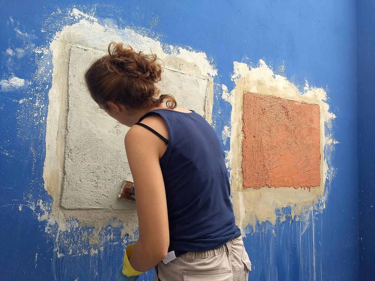 Twee proefvlakken met natuurlijke pleister op een blauwe muur, een vrouw strijkt het linkse glad